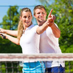 Para uśmiechniętych ludzi na korcie tenisowym, mężczyzna pokazuje kobiecie, jak trzymać rakietę.